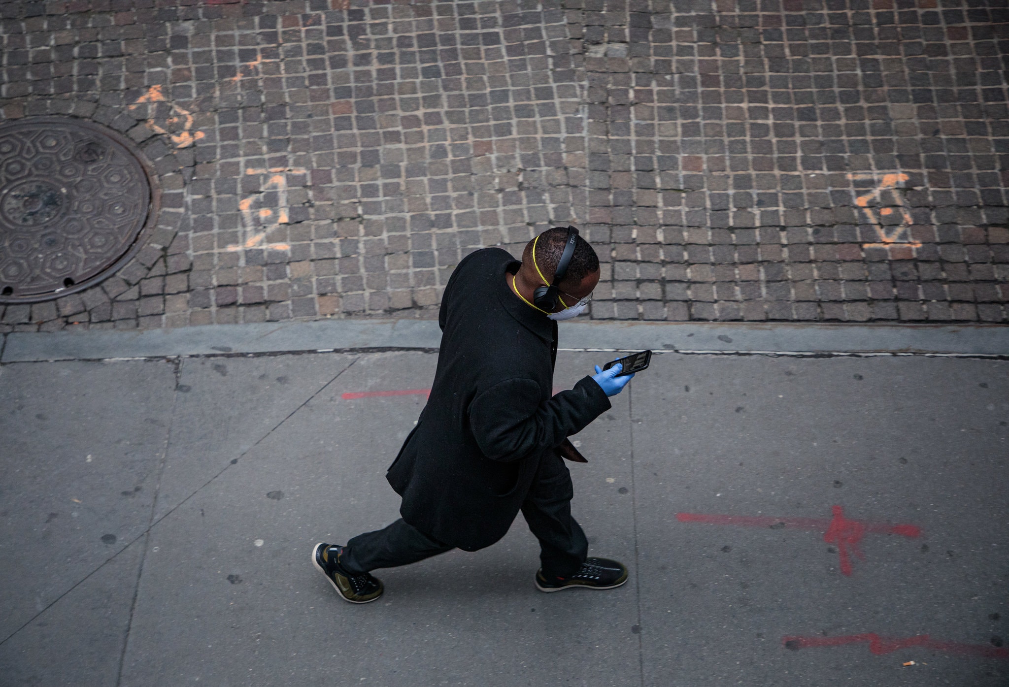 Birdseye view of a man walking down a street wearing a black coat, sneakers, headphones, a protective face mask and hospital gloves looking at his cell phone.