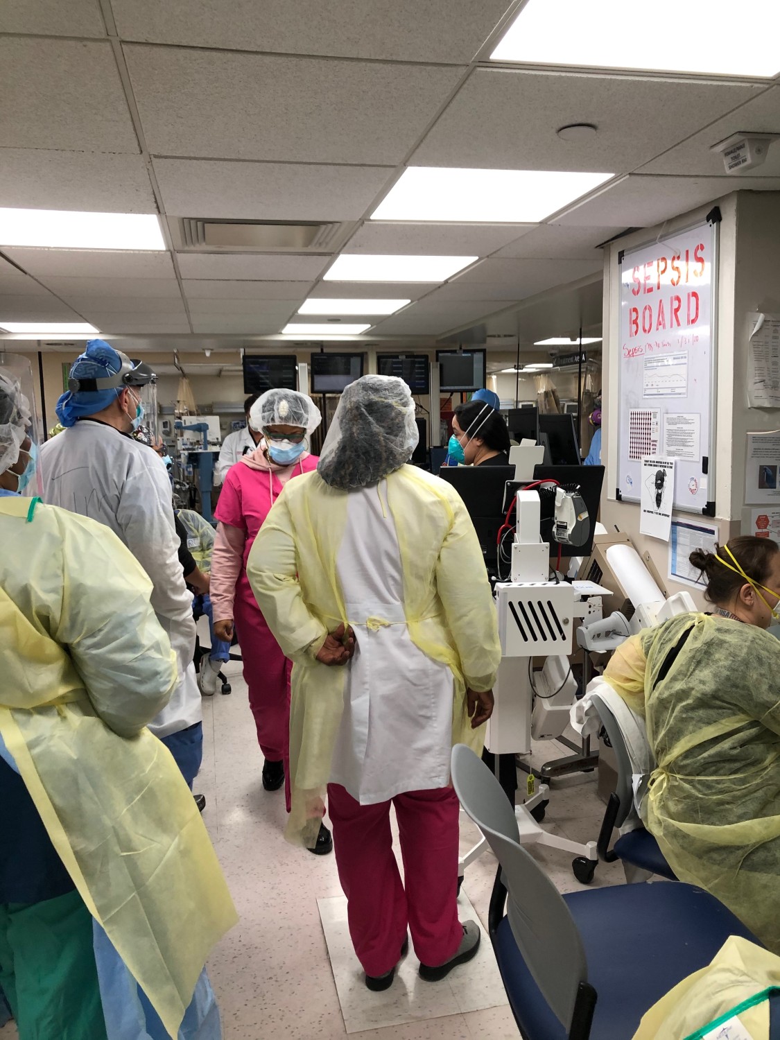 Healthcare workers in full PPE gaze at the "Sepsis Board" in the Emergency Room.