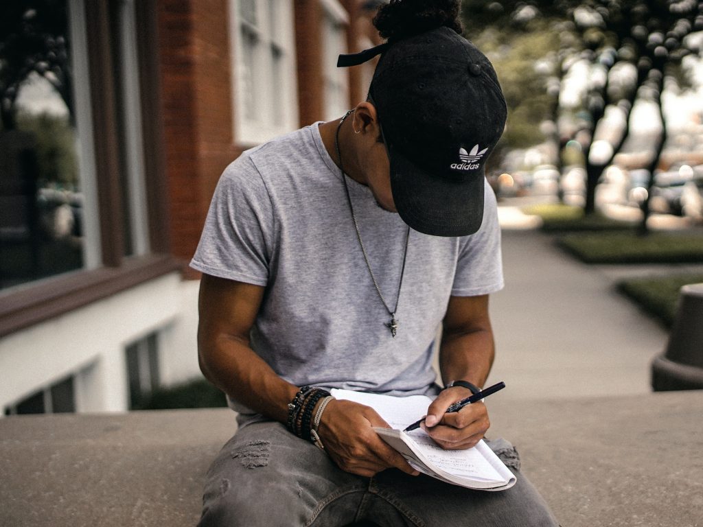 Young male journalist sitting outside on a city bench writing in a notebook.