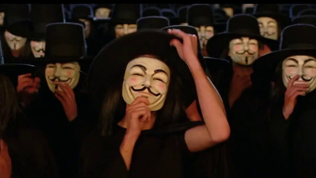 Several people at night wearing black hoods and clothing and "Anonymous" masks.