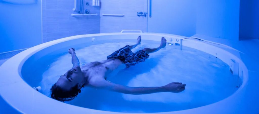 Dr. Justin Feinstein article: youn man in boxer shorts style bathing suit floats in a therapeutic floating pool in a blue-hue lit room.