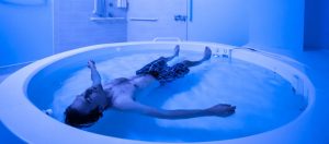 Dr. Justin Feinstein article: youn man in boxer shorts style bathing suit floats in a therapeutic floating pool in a blue-hue lit room.
