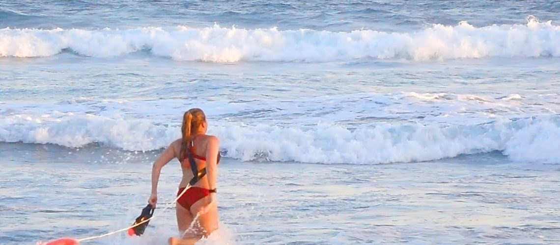 Female lifeguard in red bathing suit with two life buoys runs into the ocean beach water