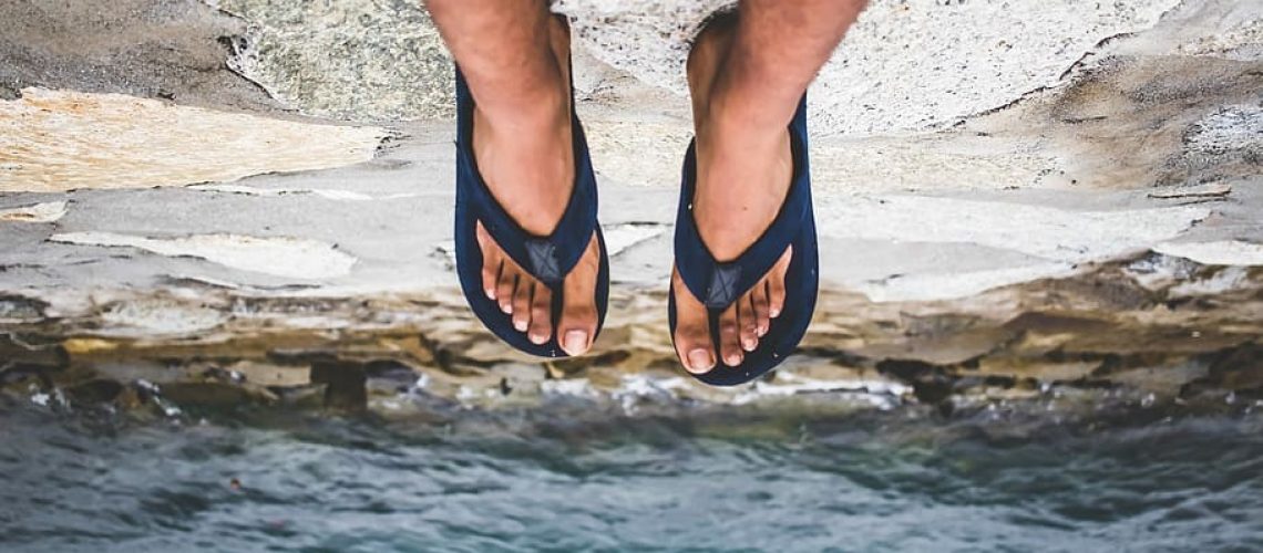 Legs and feet of a dark-skinned person in black flip flops seen dangling over a cliff with water below.
